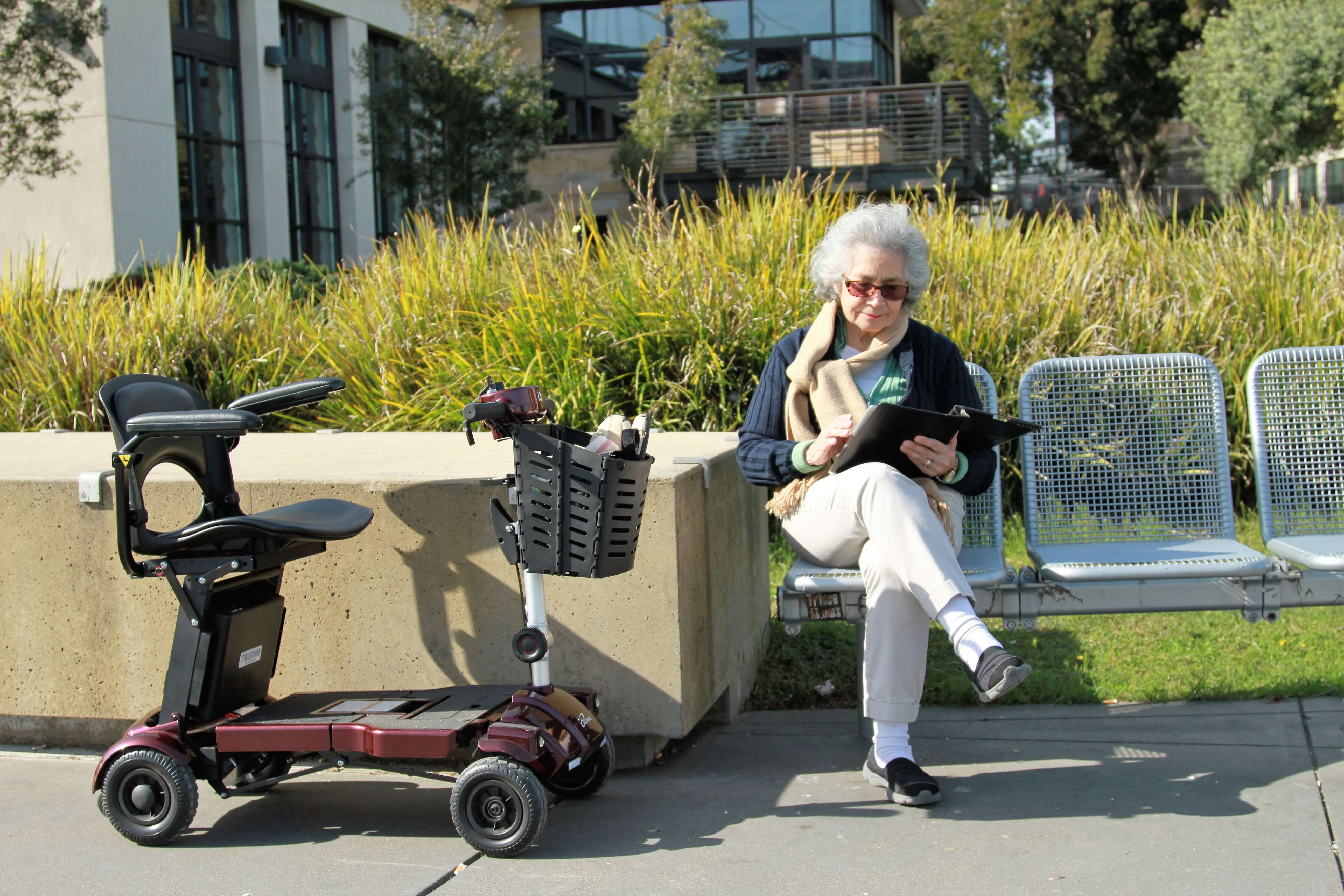 A woman relaxing next to her mobility scooter, reading a book on a park bench